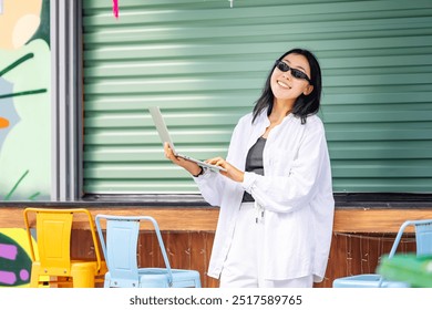 A young Asian woman wearing glasses and a white shirt types on her laptop while standing outside a colorful cafe. She appears focused, working remotely, with vibrant decor in the background. - Powered by Shutterstock