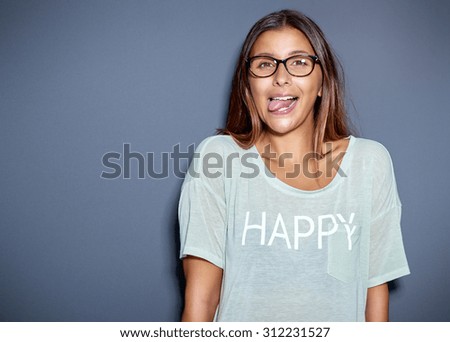 Young Asian woman wearing glasses and a casual t-shirt sticking out her tongue at the camera in a playful or rude gesture, over grey with copyspace