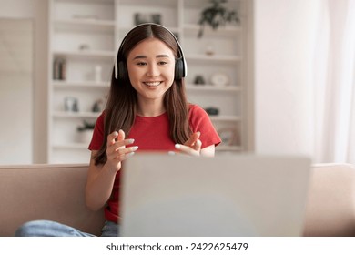 Young asian woman wearing headphones engaging in video call on laptop, smiling korean female gesturing with her hands in front of computer in bright cozy living room, enjoying online communication - Powered by Shutterstock