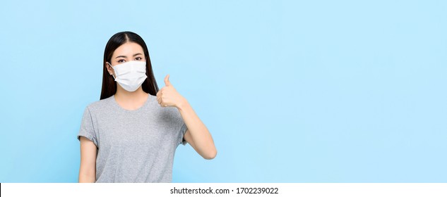 Young Asian Woman Wearing Medical Face Mask And Giving Thumbs Up Isolated On Light Blue Banner Background With Copy Space