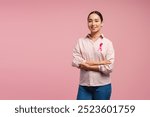 Young Asian woman wearing pink shirt with pink ribbon showing support for breast cancer awareness on pink background. Health care, support concept