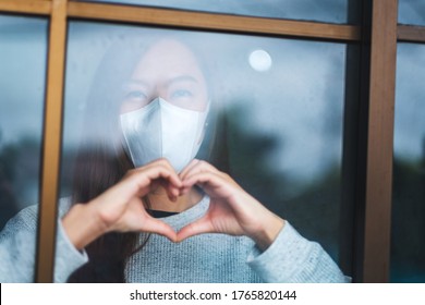 A young asian woman wearing protective mask doing heart hand sign while staying isolation at home for self quarantine and COVID-19 concept 