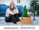 Young asian woman wearing santa hat sitting on floor with christmas trees, feeling stressed and tired during christmas holidays preparation at home