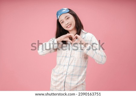 Young Asian woman wearing sleep mask and pajama smiling in love doing heart symbol shape with hands isolated on pink background. romantic concept.