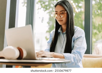 Young asian woman wearing stylish eyeglasses works on her laptop in a cozy cafe, exuding confidence and cheerfulness. The modern setup reflects a blend of technology and comfort - Powered by Shutterstock