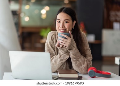 Young Asian woman wearing a sweater sitting and drink coffee at the office work equipment is placed at the table. Looking at camera. - Powered by Shutterstock