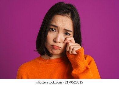Young asian woman wearing sweater crying while wiping her tears isolated over purple background
