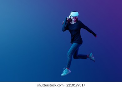 Young Asian woman wearing VR headset with experience playing video game and jumping levitating in the air on futuristic purple cyberpunk neon light background. Metaverse technology concept. - Shutterstock ID 2210199317