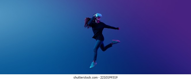Young Asian woman wearing VR headset with experience playing video game and jumping levitating in the air on futuristic purple cyberpunk neon light banner background. Metaverse technology concept. - Shutterstock ID 2210199319