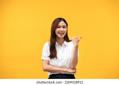 Young Asian Woman Wearing White Short Sleeve Shirt And Smiling While Using Hand And Finger To Pointing Behind Back With Crossed One Arm Isolated Over Yellow Background.