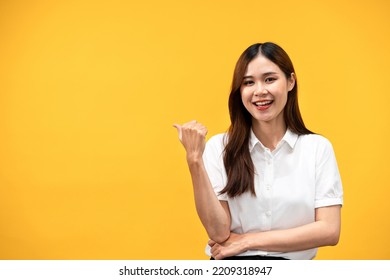 Young Asian Woman Wearing White Short Sleeve Shirt And Smiling While Using Hand And Finger To Pointing Behind Back With Crossed One Arm Isolated Over Yellow Background.