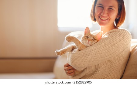 Young Asian Woman Wears Warm Sweater Resting With Tabby Cat On Sofa At Home One Autumn Day. Indoor Shot Of Amazing Lady Holding Ginger Pet. Morning Sleep Time At Home. Soft Focus.