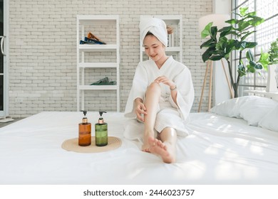 Young asian woman in a white bathrobe and towel on head, sit on a bed with lotion bottle nearby. Spreading body lotion cream gel on leg and knee after showering and taking bath, healthcare - Powered by Shutterstock