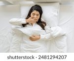 Young asian woman in white pajama use her hand to cover her mouth while yawning, lying on bed with soft pillow and a comfortable duvet wrapped around. Top view