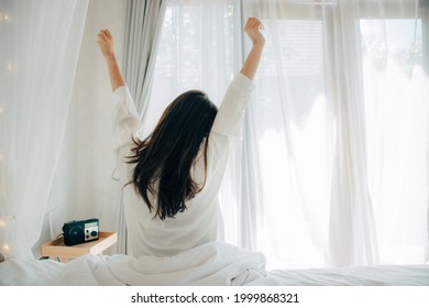 Young Asian Woman In White Pajamas Waking Up In Her Bed Sitting Near The Big Window Fully Rested And Open The Curtains In The Morning To Get Fresh Air And Sunshine. Happy Young Girl Greets New Day.