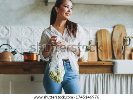 Young asian woman in white shirt hold knitted rag bag shop with green apples in hands on kitchen