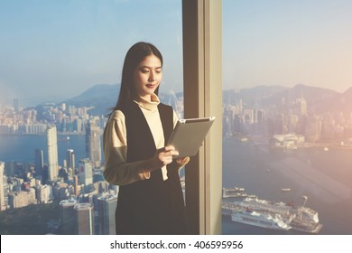 Young Asian Woman Worker Of Successful Law Firm Is Reading E-mail In Internet Via Digital Tablet From Her Client, While Is Standing In Modern Interior Against Skyscraper Window With New York City View