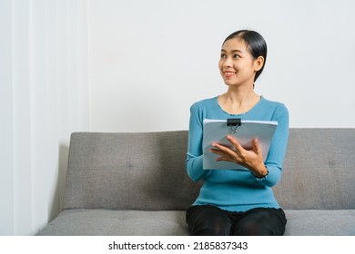 Young Asian Woman Working With Document Paper Files When Sitting On Couch, Lifting A Paper From A Stack.