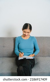 Young Asian Woman Working With Document Paper Files When Sitting On Couch, Lifting A Paper From A Stack.