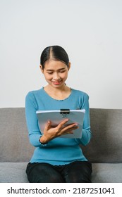 Young Asian Woman Working With Document Paper Files When Sitting On Couch, Lifting A Paper From A Stack.