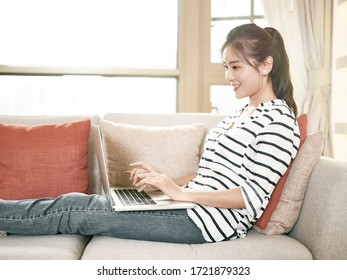 Young Asian Woman Working From Home Sitting On Couch Using Laptop Computer