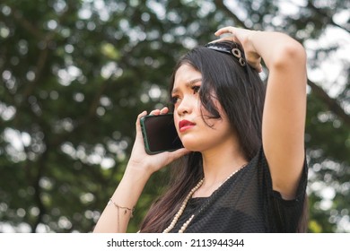 A Young Asian Woman Is Worried After Receiving Threats On Her Cellphone. Anxious Lady Listening To A Threatening Phone Call While At The Park.