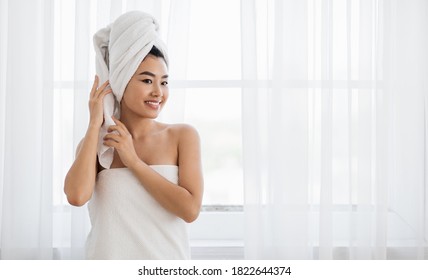 Young asian woman wrapped in towel standing next to window at bedroom, drying her hair after morning shower. Smiling lady with towel on her head looking at empty space, home interior, panorama - Powered by Shutterstock