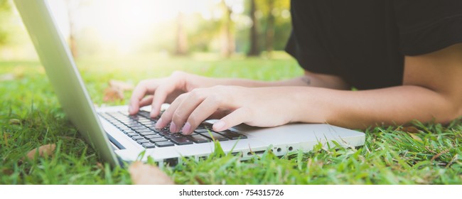 Young Asian Woman's Legs On The Green Grass With Open Laptop. Girl's Hands On Keyboard. Distance Learning Concept. Happy Hipster Young Asian Woman Working On Laptop In Park. Panoramic Banner.