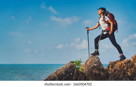 Young Asian Women Hikers Climbing Up On The Peak Of Mountain Near Ocean. Woman Hiking In The Mountains Standing On A Rocky Summit Ridge With Backpack And Pole Looking Out Over Ocean Landscape