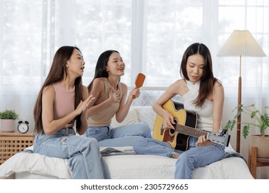 young Asian women showcasing aesthetic enjoy a music session in university dormitory. woman is playing an acoustic guitar while the other sits on the bed using a hairbrush as a makeshift microphone