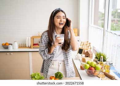 Young Asian Women Talk On Mobile Smartphone And Smile Inside Of The Kitchen In The Morning While Eating Breakfast. Telecommunication Wireless Voice Technology In People Lifestyle.