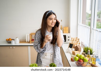 Young Asian Women Talk On Mobile Smartphone And Smile Inside Of The Kitchen In The Morning While Eating Breakfast. Telecommunication Wireless Voice Technology In People Lifestyle.