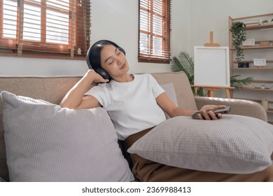 Young Asians woman listen to music on couch in living room at home. Happy Asia female using mobile smartphone, wearing headset and sitting on sofa - Shutterstock ID 2369988413