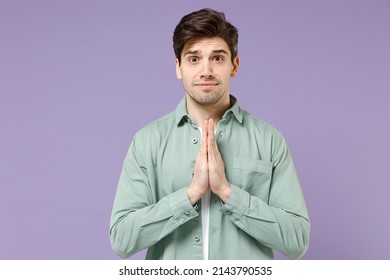 Young Asking Caucasian Man 20s Wearing Mint Shirt White T-shirt Hold Hands Folded In Prayer Gesture, Begging About Something Isolated On Purple Background Studio Portrait. People Lifestyle Concept.