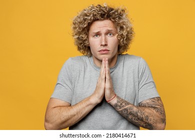 Young Asking Caucasian Man 20s He Wear Grey T-shirt Hold Hands Folded In Prayer Gesture Begging About Something Making Wish Isolated On Plain Yellow Backround Studio Portrait. People Lifestyle Concept