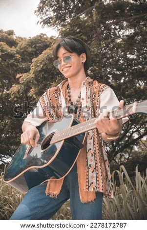 A young aspiring asian guitarist with his old guitar at a nature park or indie concert. Folk rock enthusiast.