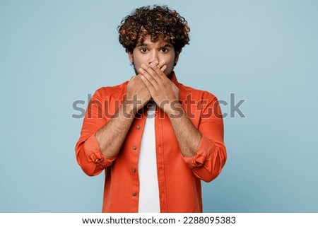 Young astonished shocked sad Indian man wear orange red shirt white t-shirt cover mouth with hands look camera isolated on plain pastel light blue cyan background studio portrait. Lifestyle concept