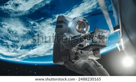 Young Astronaut Floating in Space in Zero Gravity. Spaceman Using Laptop Computer in Open Space Outside an Orbiting Spacecraft, Browsing Instruction Manuals for Maintenance Work on a Satellite