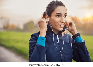 Young athlete adjusting jacket while listening to music at park. Smiling young woman feeling relaxed after a long run during the sunset. Happy sporty woman smiling and looking away during workout. - Powered by Shutterstock