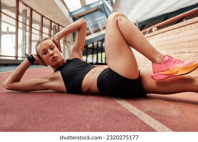 A young athlete girl is warming up before training at the stadium. A slender blonde woman in a black tracksuit kneads her muscles and does exercises for the abdominals. Sports and recreation. - Powered by Shutterstock