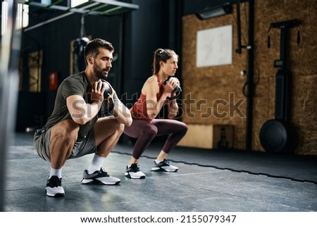 Young athlete using kettlebells while exercising squats during sports training in health club. Focus is on man.