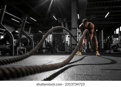 young athletic african american man trains with ropes in dark gym, motivated guy waving ropes in fitness club - Powered by Shutterstock