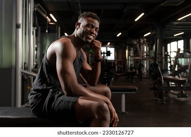 young athletic african american man sits in dark gym and smiles, athletic guy rests and looks at the camera in fitness club, portrait of athlete in training - Powered by Shutterstock