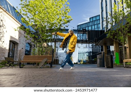 Young And Athletic B-Boy Breakdancing To Break Beats On The City Street Among Modern Buildings Alone With A Speaker. Fashionable Man Practicing Freestyle Choreography For Upcoming Break Dance Battle.