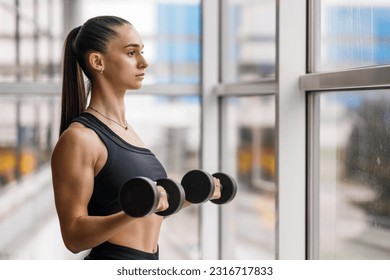 Young athletic fitness girl doing biceps curls with dumbbells near the window.