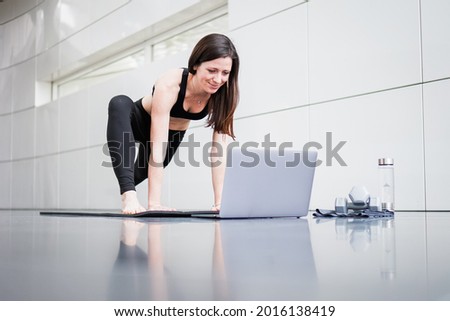 Young athletic fitness woman watching online stretching training video on yoga mat in triangle pose infront of a laptop at home studio indoor with panoramic window on reflective modern background