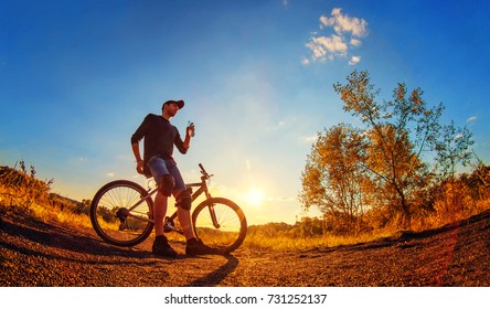 Young Athletic Guy In A Black T-shirt, Blue Jeans Shorts And Knee Pads On A Sports Bike Holds In Hand A Plastic Bottle With Water On An Colorful Autumn Background  In Sunset Light.