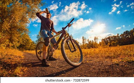 Young Athletic Guy In A Black T-shirt And Baseball Cap On A Sports Bike Drinks Water From A Shaker On An Autumn Background With A Bright Sun In The Sky.