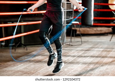 Young Athletic Man In Black Uniform Training With A Jumping Rope, Warming Up On The Boxing Ring In The Gym. Croppped Image With No Face