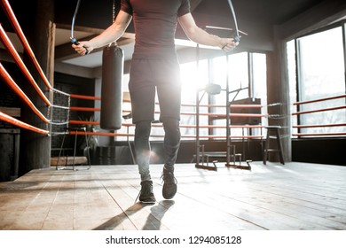 Young Athletic Man In Black Uniform Training With A Jumping Rope, Warming Up On The Boxing Ring In The Gym. Croppped Image With No Face